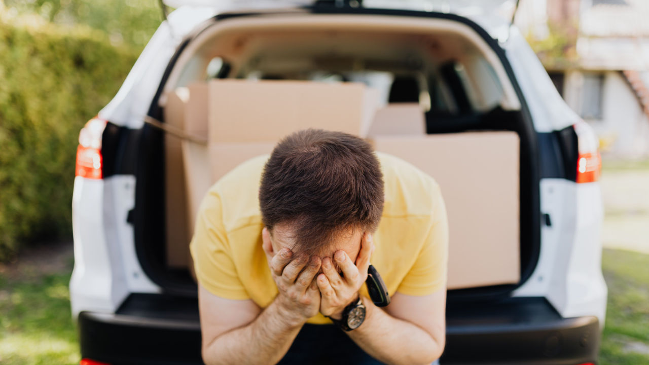 Man Holds His Head In Despair With Car Boot Crammed Full of Boxes Behind Him
